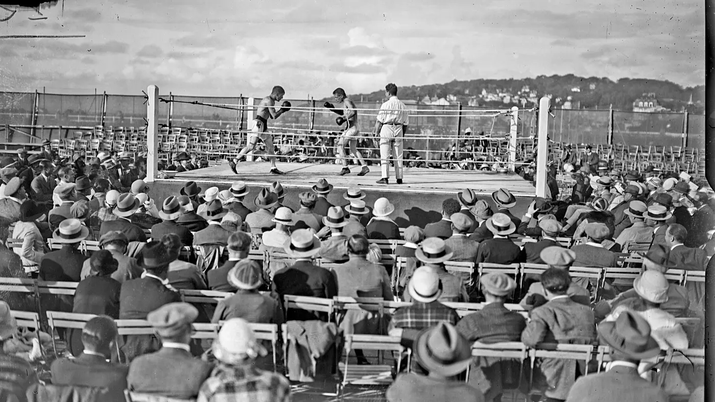 A crowd of spectators in straw hats watching an open-air boxing bout in 1924