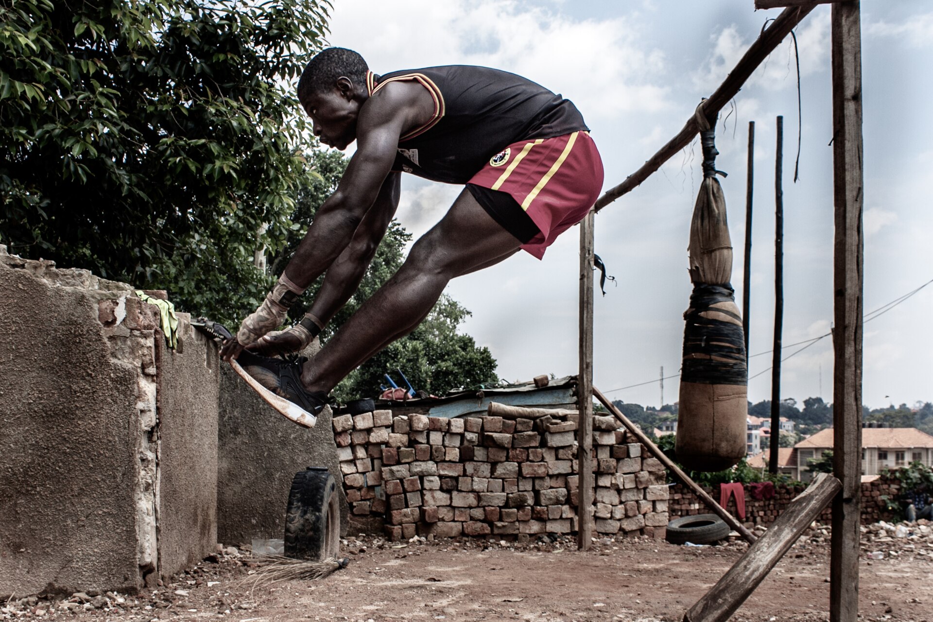 An athlete performs a mid-air jump-tuck exercise in an outdoor training area beside a makeshift punching bag