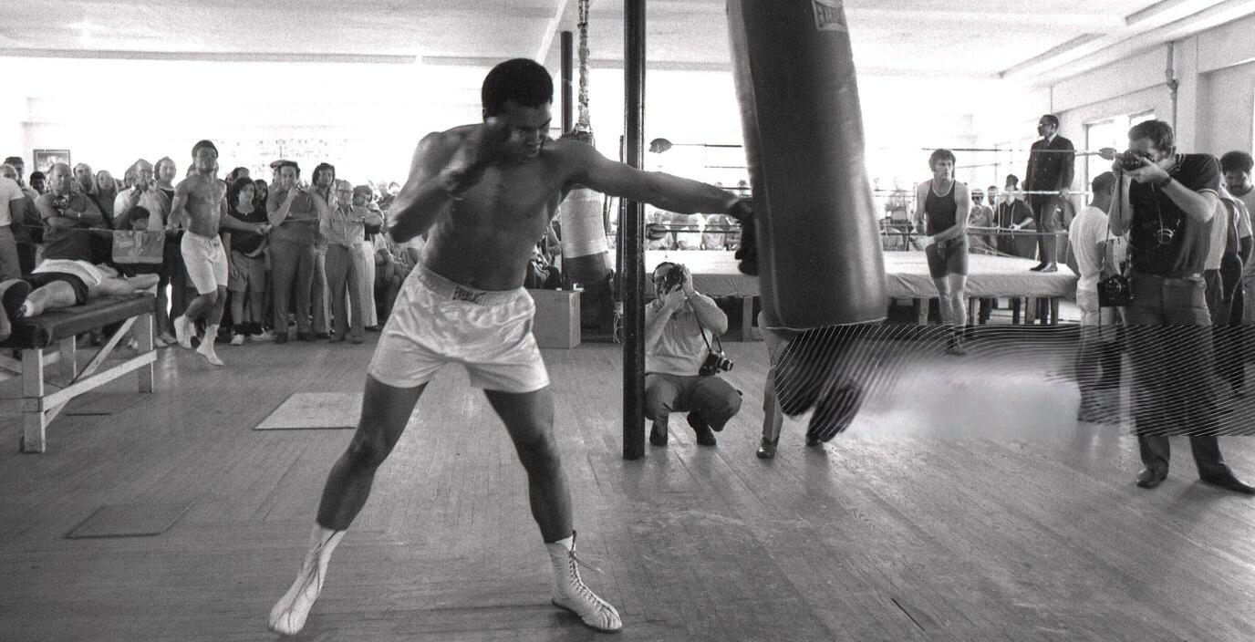 Muhammad Ali punches a heavy bag in a crowded gym while photographers capture the moment