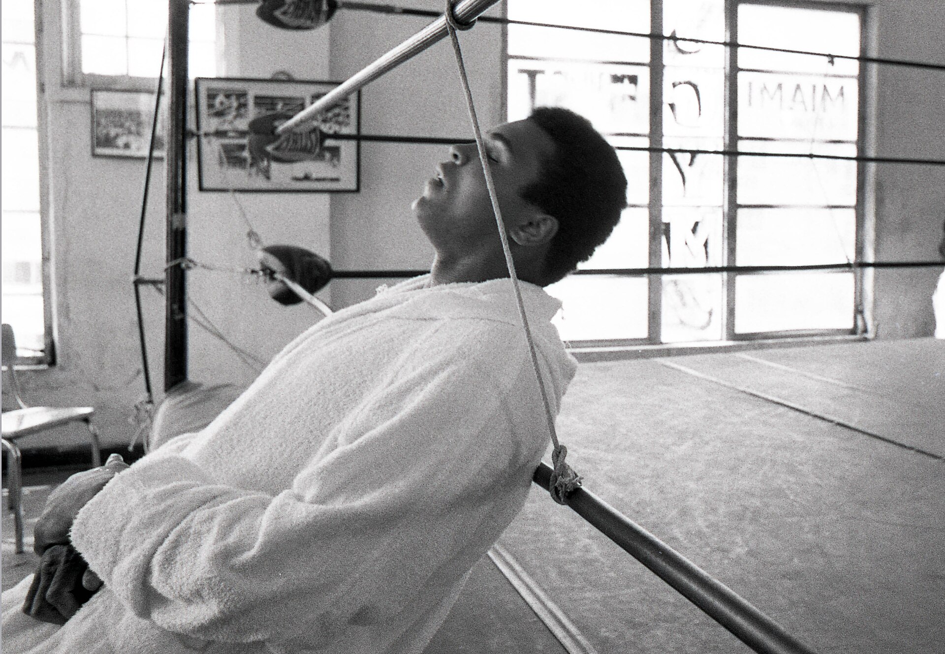 A black and white photograph of a boxer resting his head against the ropes in a boxing ring