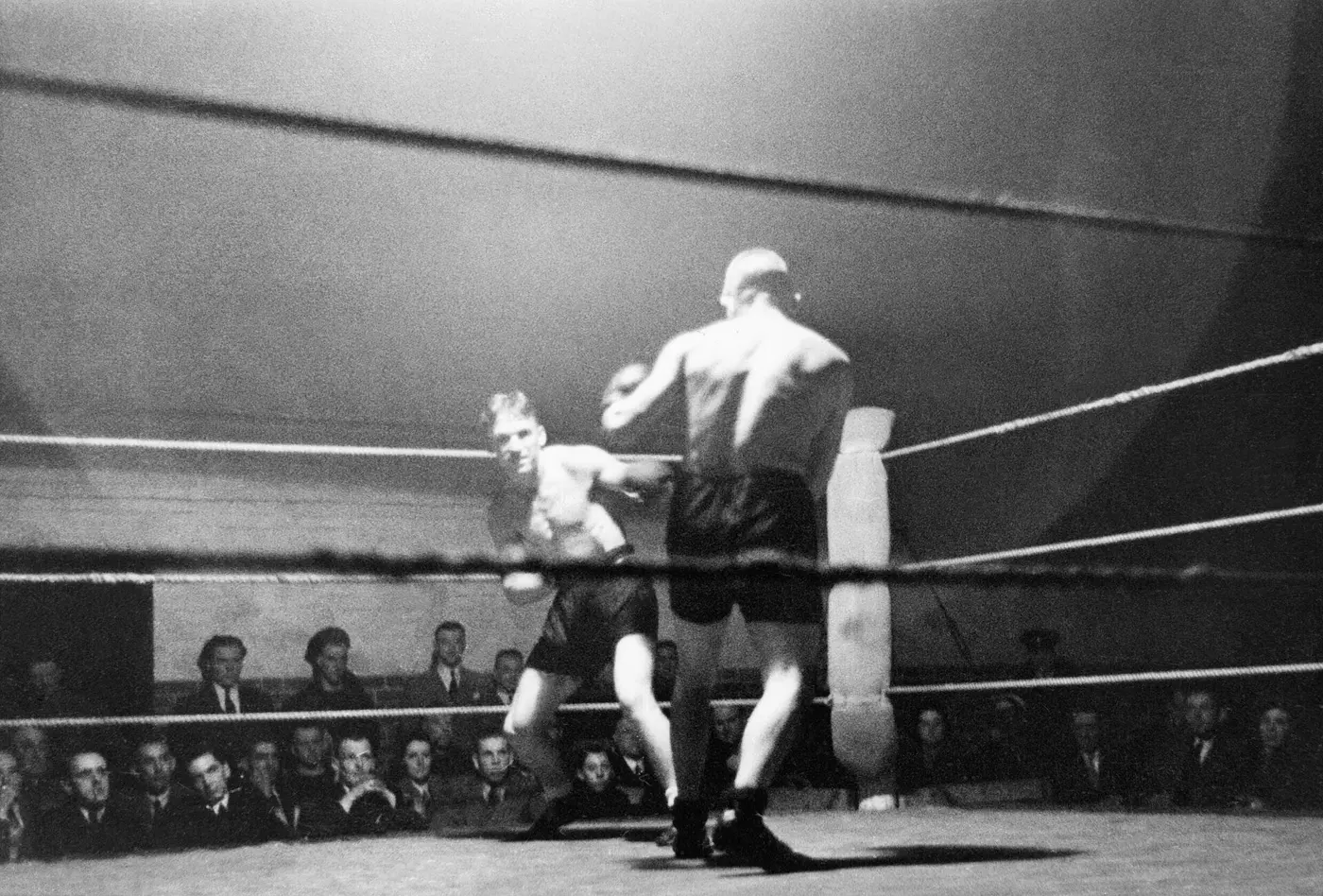 A mid-punch moment at a wartime charity boxing bout, seated crowd in the background
