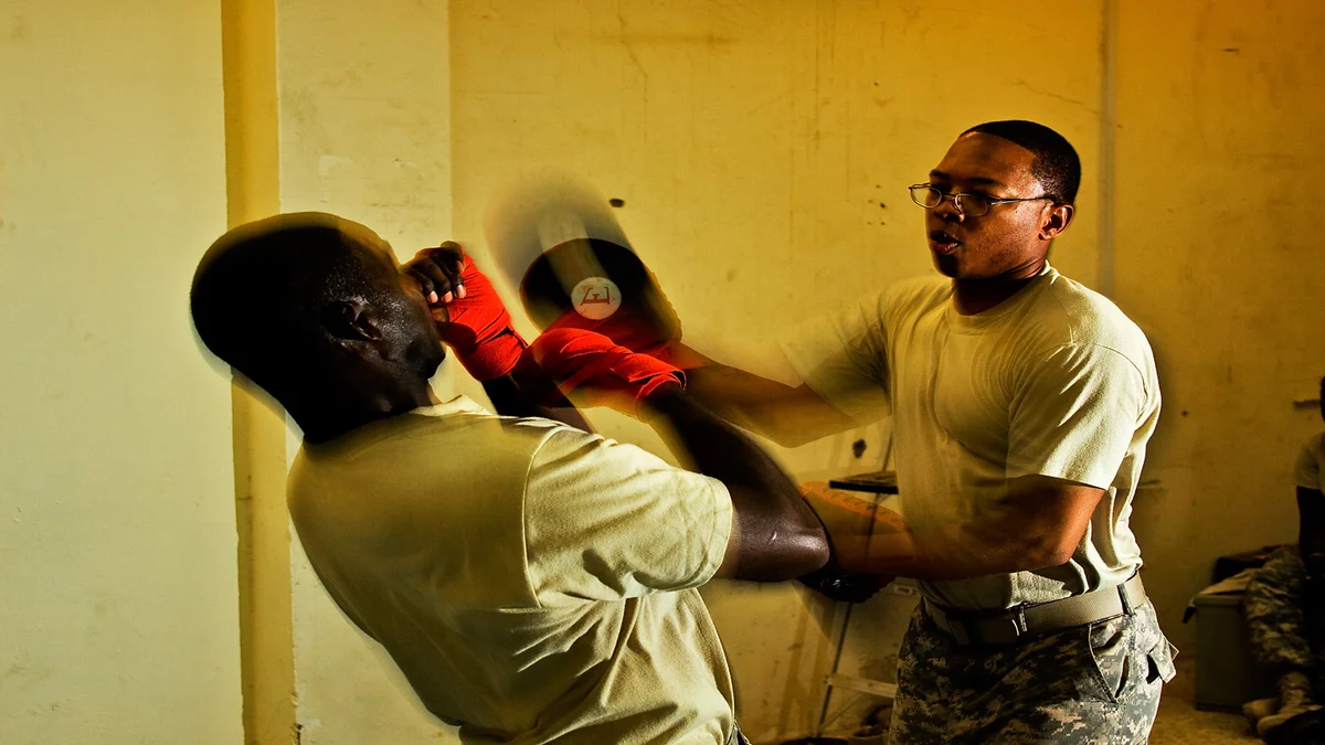 Two men in military uniform training with wrapped hands and focus mitts indoors, one throwing a punch
