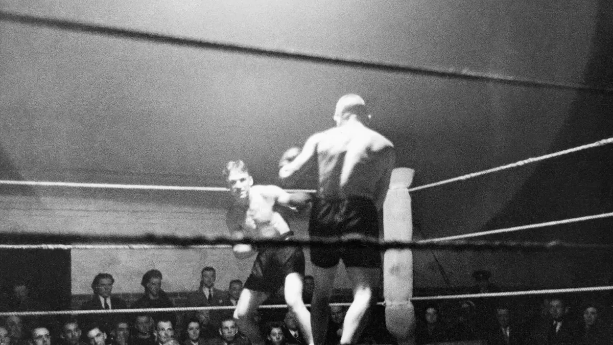Two boxers mid-exchange in a roped ring, watched by a seated crowd, mid twentieth century