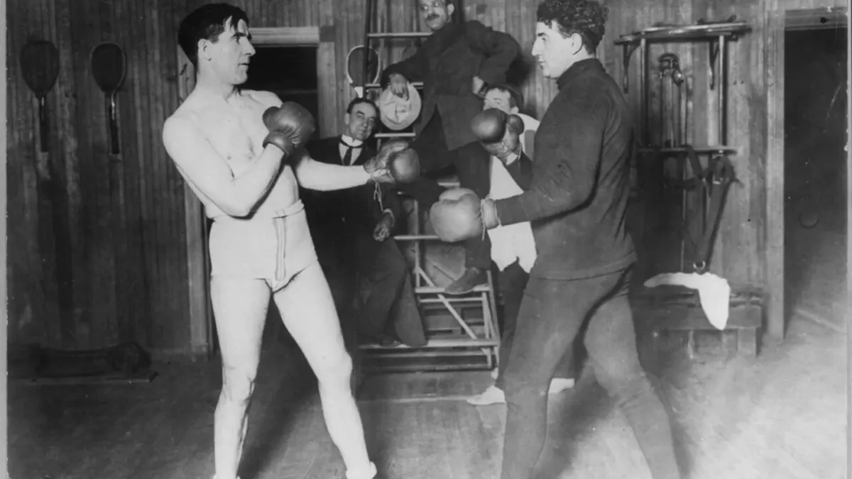 Two boxers sparring in a gym in the early 1900s, watched by men in suits