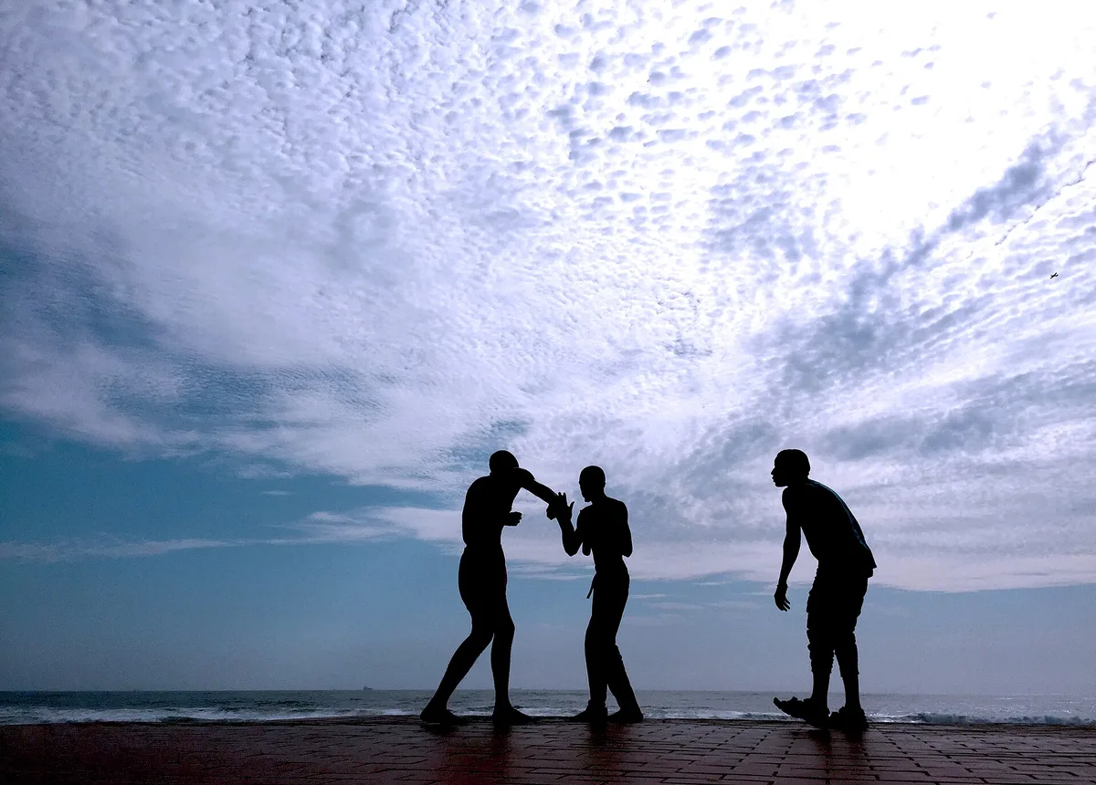 Two silhouetted figures shadow boxing outdoors at dusk while a third looks on